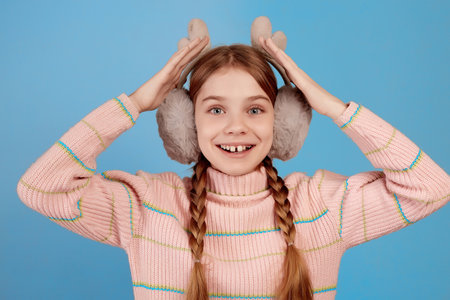 Little surprised girl wearing fluffy headphones on blue background. Close portrait.の写真素材