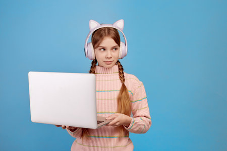 Little girl with headphones on her head and laptop looking aside on blue background.の写真素材