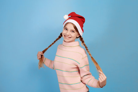 Little smiling girl wearing Santa hat holding pigtails on blue background.の写真素材