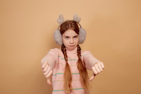 Little upset girl with fluffy headphones showing thumb down on beige background.の写真素材