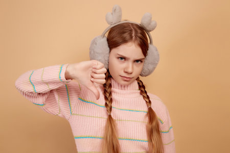 Little dissatisfied girl with fluffy headphones showing thumb down on beige background.の写真素材
