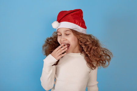 Yawning little African-American girl with Santa hat on blue background.の写真素材
