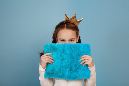 Little African-American girl with crown and notebook in hands closing her face on blue background.の写真素材