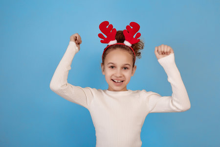 Happy little African-American girl with decorative deer horns with hands up on blue background.の写真素材