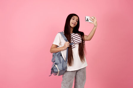 Young smiling teenage girl taking a selfie with phone with bag on pink background in studio.の写真素材