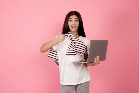 Young smiling teenage girl and laptop shows thumb up on pink background in studio.の写真素材