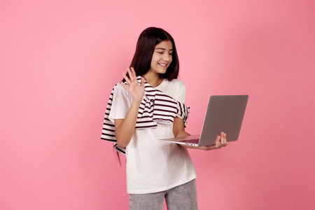 Young smiling teenage girl waving on call with laptop on pink background in studio.の写真素材