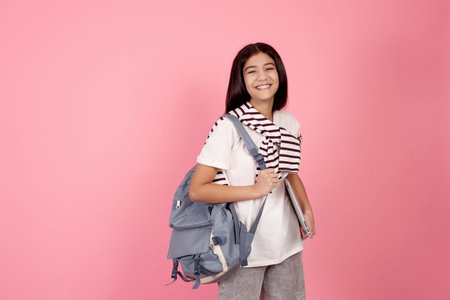 Young smiling teenage girl with bag and laptop on pink background in studio.
