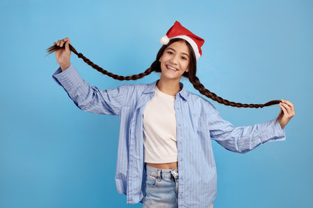 Smiling teenage girl with and Santa hat holding pigtails on blue background.の写真素材
