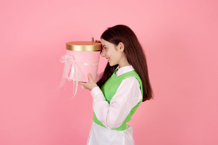 Smiling teenage girl holding present box and looking inside on pink background in studio.