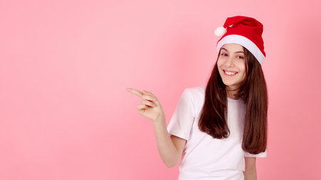 Smiling teenage girl with Santa hat showing aside by fingers on pink background in studio.の写真素材