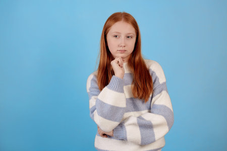 Thinking redhead teenage girl on blue background in studio.