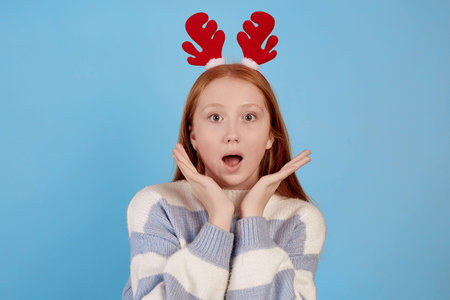 Surprised redhead teenage girl with decorative deer horns on blue background.