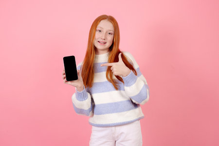 Smiling redhead teenage girl wearing sweater holding phone showing screen on pink background in studio.の写真素材