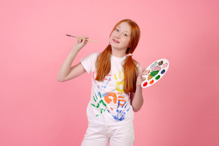 Happy and cheerful redhead teenage girl with paints and brush smiling on pink background in studio.の写真素材