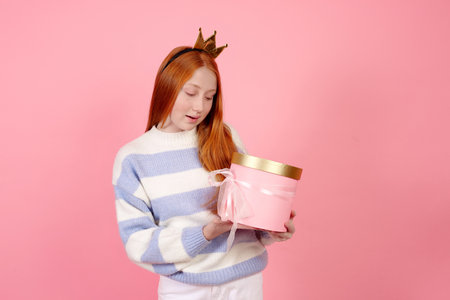 Smiling redhead teenage girl wearing crown and sweater holding present box on pink background in studio.の写真素材