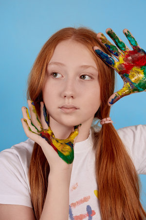 Happy and cheerful ginger teenage girl showing painted hands on blue background in studio.の写真素材