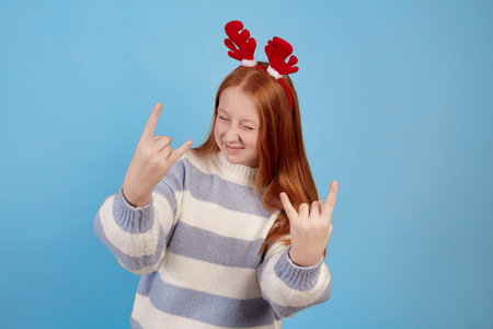Smiling redhead teenage girl with decorative deer horns dancing and showing sign of the horns on blue background in studio.の写真素材