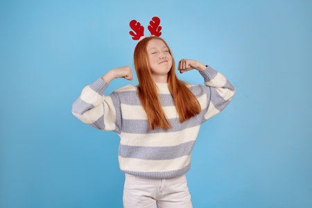 Smiling redhead teenage girl with decorative deer horns raises arms and shows biceps on blue background in studio.の写真素材