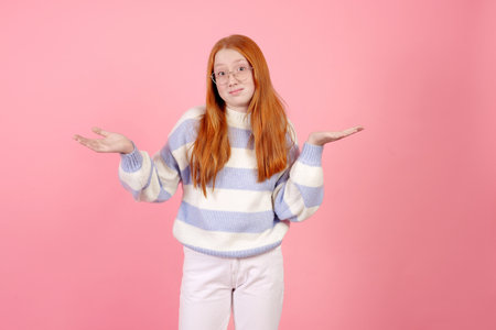 Confident redhead teenage girl wearing glasses and sweater on pink background in studio.の写真素材