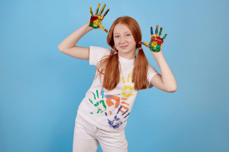 Happy and cheerful ginger teenage girl showing painted hands on blue background in studio.の写真素材