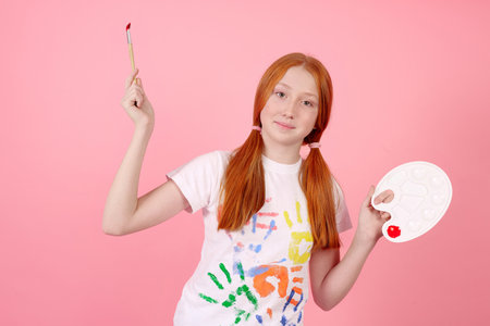 Happy and cheerful redhead teenage girl with paints and brush smiling on pink background in studio.の写真素材