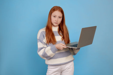 Confident redhead teenage girl holding laptop on blue background in studio.