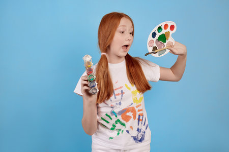 Happy and cheerful ginger teenage girl with paints and brush smiling on blue background in studio.の写真素材