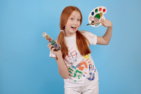 Happy and cheerful ginger teenage girl with paints and brush smiling on blue background in studio.の写真素材
