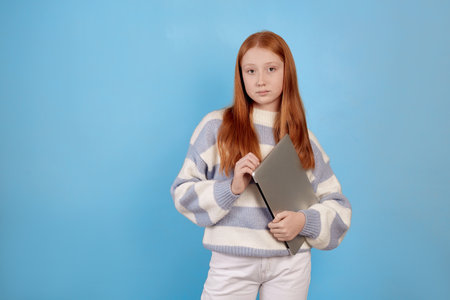 Confident redhead teenage girl holding laptop on blue background in studio.の写真素材