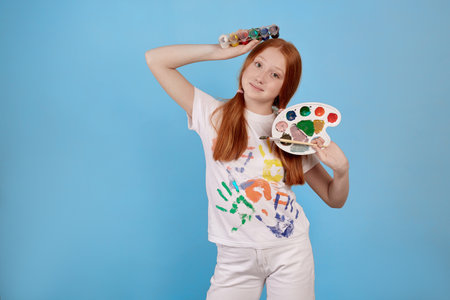 Happy and cheerful ginger teenage girl with paints and brush smiling on blue background in studio.の写真素材