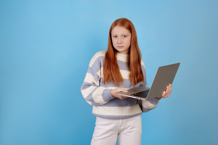 Confident redhead teenage girl holding laptop on blue background in studio.の写真素材