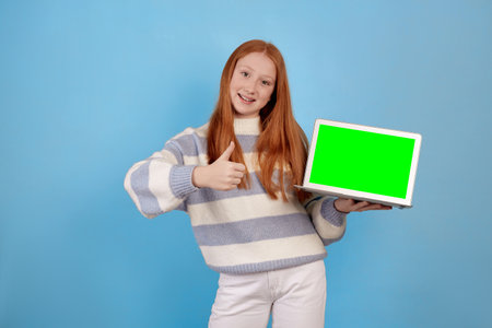 Smiling redhead teenage girl holding laptop on blue background in studio. Green screen on laptop.の写真素材