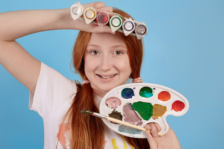 Happy and cheerful ginger teenage girl with paints and brush smiling on blue background in studio.の写真素材