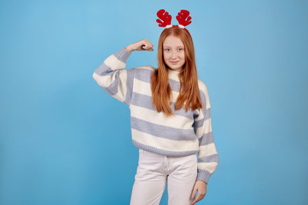 Smiling redhead teenage girl with decorative deer horns raises arms and shows biceps on blue background in studio.の写真素材
