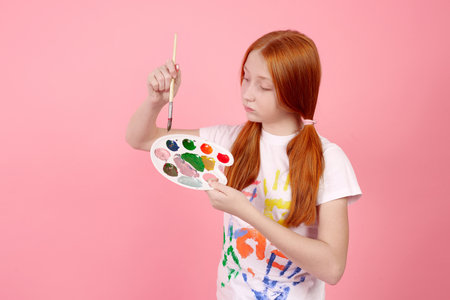 Happy and cheerful redhead teenage girl with paints and brush smiling on pink background in studio.の写真素材