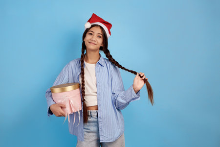 Smiling teenage girl with pigtails and Santa hat holding pink surprise box on blue background.の写真素材