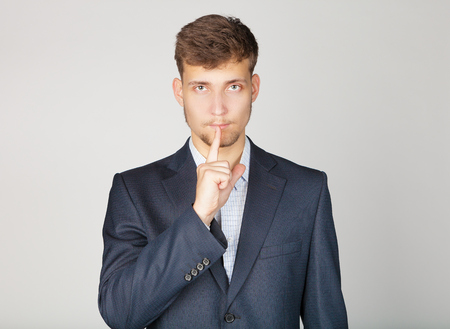 Young business man in suit stands confidently and shows the sign of silence by his finger near mouth.の写真素材