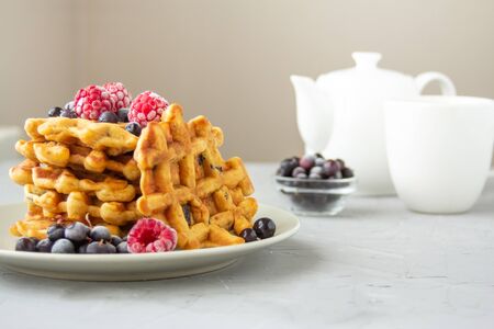 Carrot waffles with prunes, walnuts, cinnamon, ginger and nutmeg on a gray plate on a grey background. Sprinkled with fresh raspberries and black currants. Near a white teapot and a cup of teaの写真素材