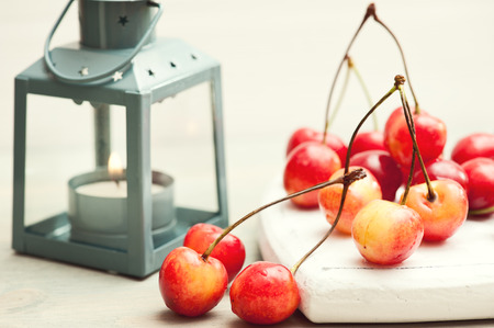 Stack of red and yellow cherries on white wooden background with lit lantern. Sepia toned imageの写真素材