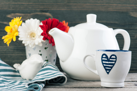 Navy blue heart cup, white teapot and colorful daisies on dark wooden background. Toned imageの写真素材