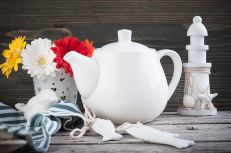 White tea pot on brown wooden table with lighthouse and colorful daisies. Place for textの写真素材