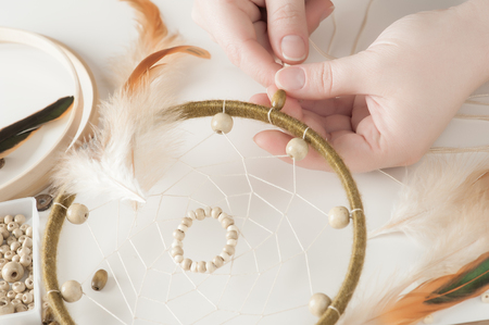 Close-up of female hands with wooden beads, having dream catcher assembled. Handmade workshop composition of feathers, beads, threads, tools for DIYの写真素材