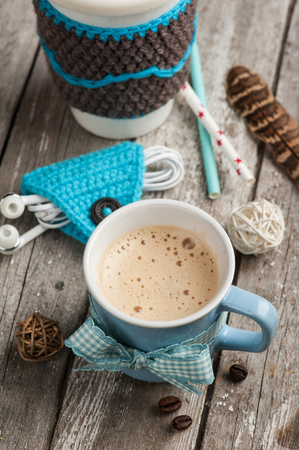 Cute blue knitted ear pods cord holder and cappuccino mug on wooden table. Life style set with cup of coffeeの写真素材
