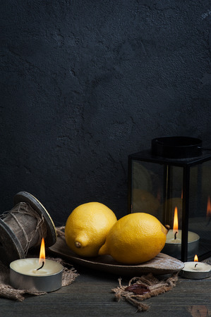 Dark still life with yellow lemons and lit candles. Textures, burlap, old wood and black concrete wall on backgroundの写真素材