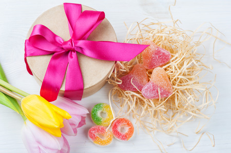 Pink yellow tulips and gift box with red ribbon, sweet hearts on a white painted wooden background. Top view with copy spaceの写真素材