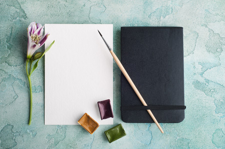 Paint brushe, purple, yellow and green watercolor paint with spring flowers and blank white paper on blue concrete background.の写真素材