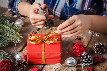 Girl sitting at her desk tying bow for Christmas present. Closeup of hands and red box with golden ribbon on dark wooden backgroundの写真素材