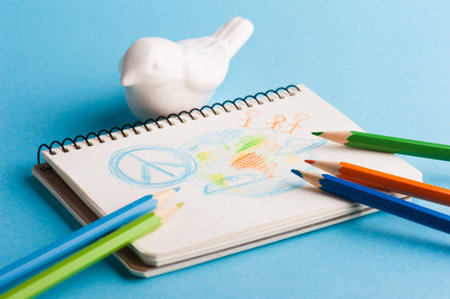 Notepad with color pencils and child drawing of peace symbol on blue background