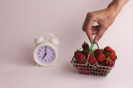 Female hand holds a mini basket with strawberries and a retro clock on a white background. Harvest time concept in the garden. Shopping in the online store. Home delivery.の写真素材